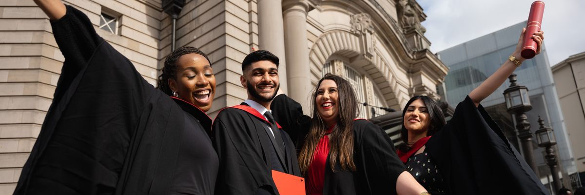 Four graduating people, 3 women and 1 man, standing outside Usher Hall with their graduation gowns, holding their degrees in their hands, being very happy.