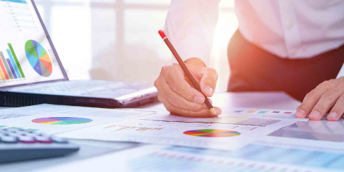 A person is looking at charts displayed on paper and on laptop and taking notes