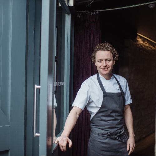 Tom Kitchin standing at the backdoor of a restaurant kitchen with an apron on