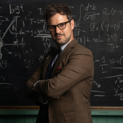 Kevin Quantum standing in front of a blackboard with his arms crossed. 