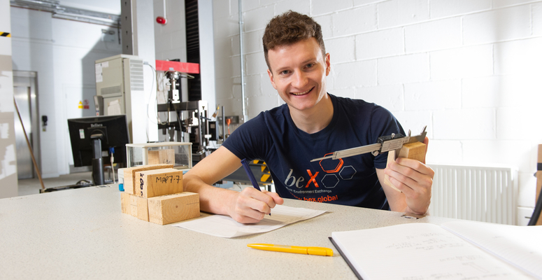 beX student Greg smiles for the camera whilst measuring wood and taking notes