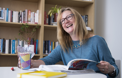 Woman laughing at a table with a book in one hand and a cup in the other hand in The Writers Room at Merchiston.