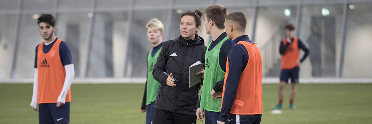 Indoor football practice with players in bibs talking to woman holding an open book