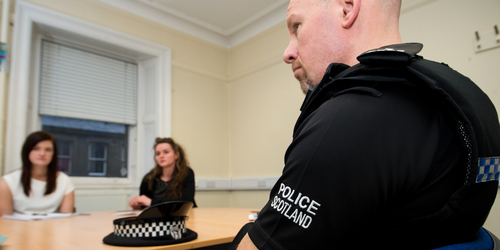 Two students, on placement at Torphicen Street Station, sitting across the desk from a policeman
