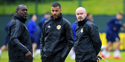 3 people in Scottish Football Association jackets stand close together on a football pitch, with players training in the background.