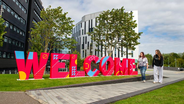 Two students walking beside a large red welcome sign outside the sighthill campus