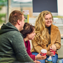 Three graduates seated together indoors, chatting over takeaway coffee cups in a informal office setting.