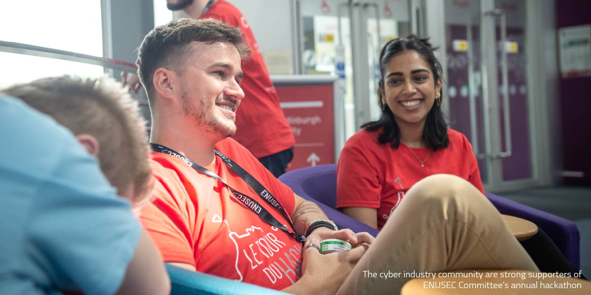 Three people sit in a casual indoor lounge area, wearing red T-shirts, engaged in conversation. One person in the foreground is seated with a lanyard and holding a drink, while another sits nearby smiling. In the background, glass doors and signage are visible. Overlay text reads: “The cyber industry community are strong supporters of ENUSEC Committee’s annual hackathon.”
