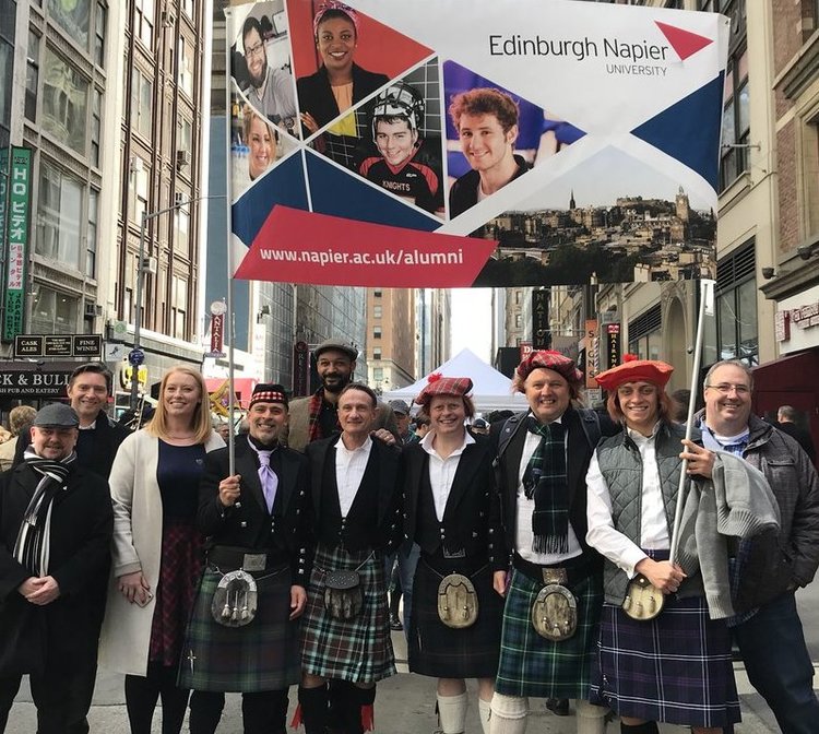 A group of people, dressed in kilts and tartan, stand in front of a banner promoting ENU