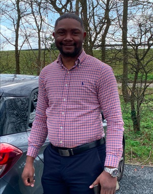 Chiemezie Nwakire standing in front of car and trees, wearing a checked shirt
