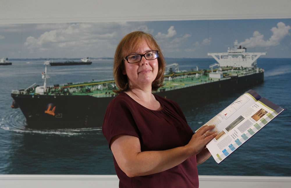 Gillian Macrosson standing in front of a large poster of a large ship, holding a book