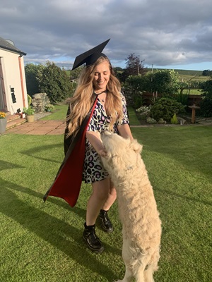 Lauren standing in garden wearing a graduation cap and gown. She's hugging her dog