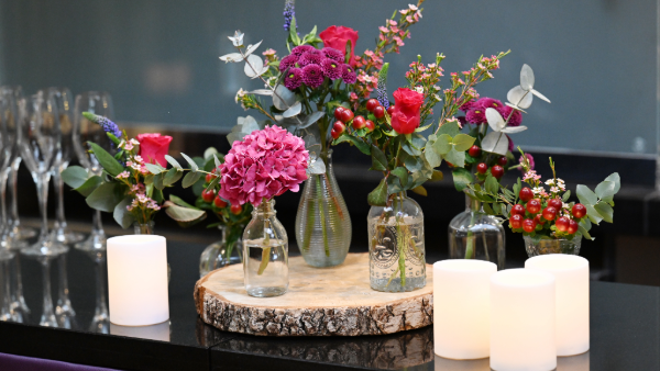 A selection of small vases containing bright flowers surrounded by drinks glasses and candles