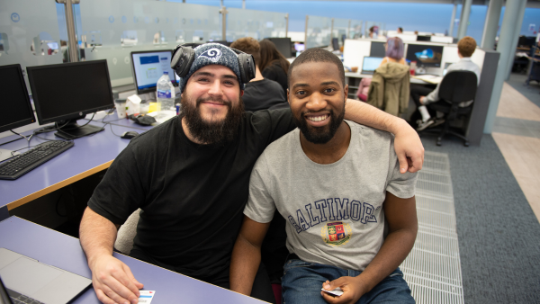 Two students sitting with their back to a computer. One student has their arm round the other. Both are smiling into the camera
