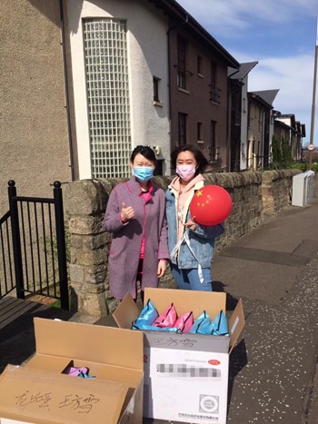 Fangxue Wang and Dixiao Shen standing on an Edinburgh street. They are wearing protective masks and holding a balloon covered in the Chinese flag. In front of them are boxes filled with protective equipment