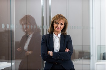 Jeanette MacIntyre wearing a dark navy suit with white shirt, standing in front of a glass wall
