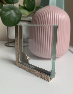 A right angle metal frame holding a square piece of glass. Placed in front of a pink vase, which can be seen through the glass, and a green plant to the left-hand side