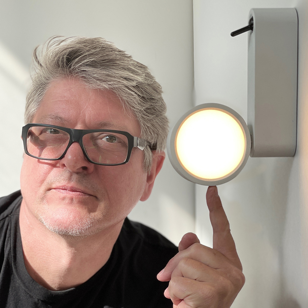Neil Poulton looking into the camera with his finger pointing towards a lamp