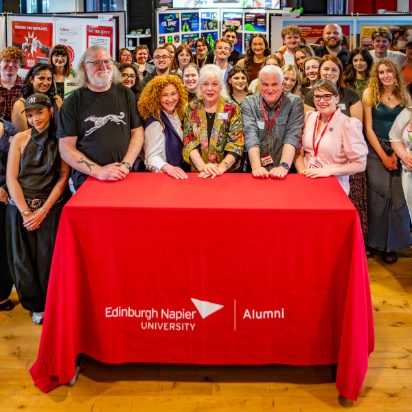 Large group gathered behind a red table at an indoor event, posing for a photo. The tablecloth reads “Edinburgh Napier University Alumni.” Around 25–30 people of mixed ages stand closely together, with five people at the front resting their hands on the table. The setting appears to be a bright room with displays on the walls in the background. Many attendees wear lanyards, suggesting an organised university or alumni gathering.