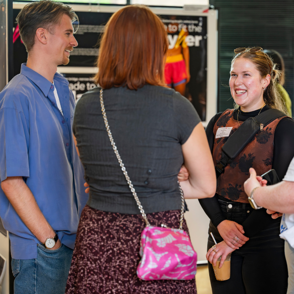 Three people chatting at an indoor event, with posters in the background and one person holding an iced coffee