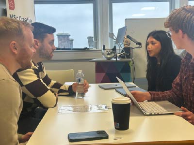 Four people sit around a table in a bright meeting room, engaged in discussion. One person uses a laptop while another holds a water bottle, and a takeaway coffee cup and smartphone are placed on the table. A microphone on a stand and audio equipment are visible near a window in the background.