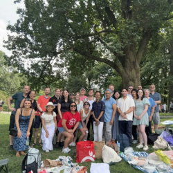 Group of alumni standing together in a park in NYC, in front of picnic blankets and bags