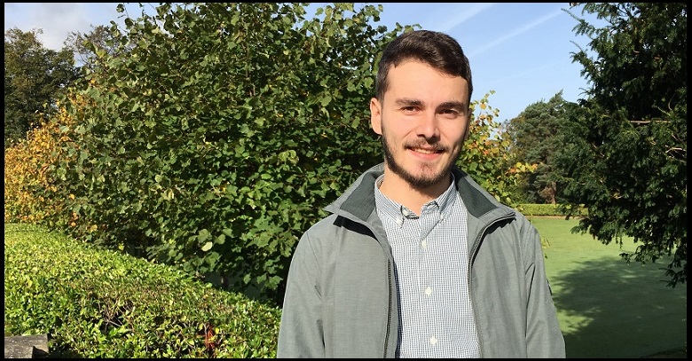 Kiril, Hospitality Management student, standing in a park with trees and bushes on a sunny spring day