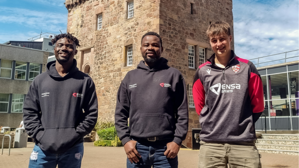 Three people standing outdoors in front of a historic stone building on a sunny day, wearing branded hoodies and sportswear.