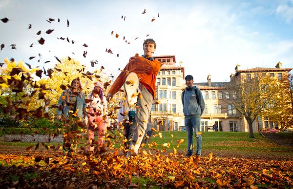 A student kicks up some autumn leaves while their friends smile in the background of Edinburgh Napier's Craiglockhart campus. 