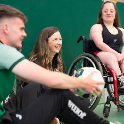 Three people taking part in an inclusive sports activity, including a wheelchair user holding a ball in an indoor gym.