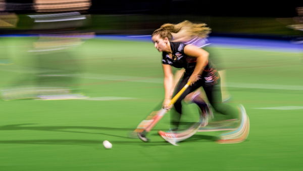Field hockey player sprinting with the ball, captured with motion blur on a green pitch.