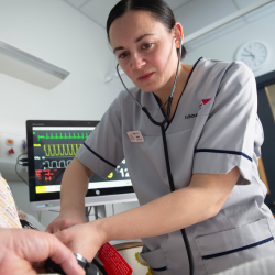 Healthcare professional using a stethoscope to examine a patient, with a heart monitor displaying vital signs in the background.