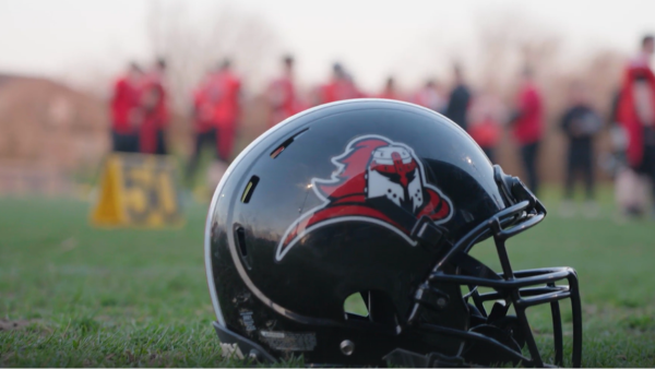 Black football helmet with a red-and-white mascot logo resting on grass, with players blurred in the background.