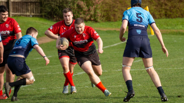 Rugby player in a red jersey runs with the ball while opponents move in to tackle on a grassy field.