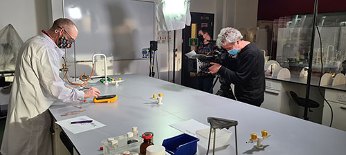 Dr McGill carries out science experiment in front of two camera operators in lab.