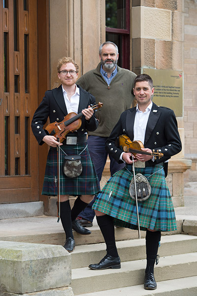 Scottish guys in kilts and holding violins in front of a building