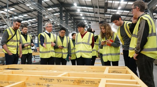 A group of students in high vis vests listening to a man speaking in a warehouse