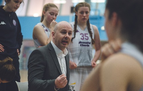 Donnie MacDonald in suit jacket and white shirt in demonstrative pose on court at the centre of a group of female basketball players
