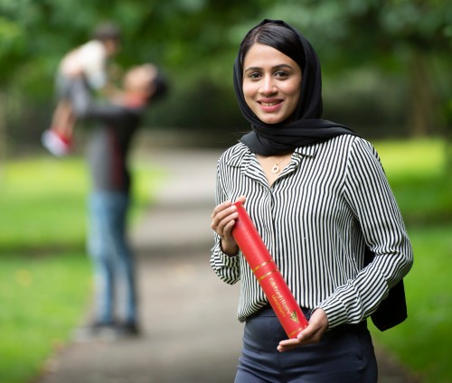 Jeshreena Palakkal in park with graduation scroll, husband and son out of focus in the background