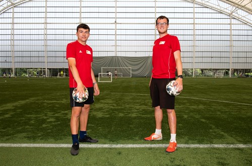 Two footballers, each holding a ball, on artificial surface under dome structure