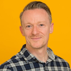 Head and shoulders of Rich Macfarlane, wearing check shirt and against a yellow backdrop