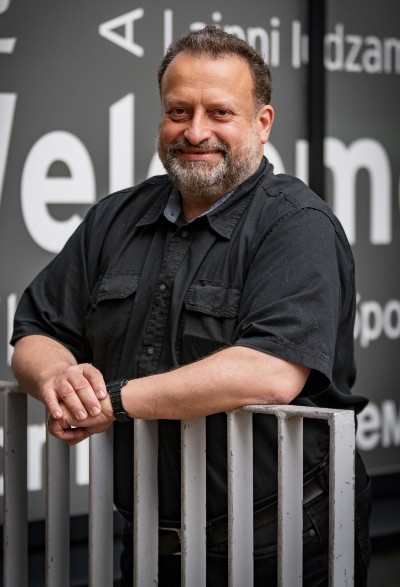 Peter Andras, bearded, on campus and leaning on a fence in front of a welcome sign