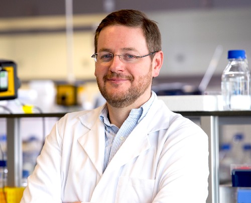 Portrait of Peter Barlow, bespectacled, bearded, in lab coat in lab setting
