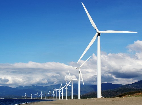 Wind turbines stretched along a coastline against a cloudy blue sky backdrop