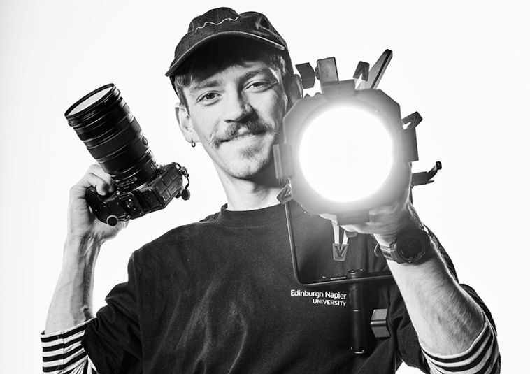 A smiling person holding a camera and a camera light.