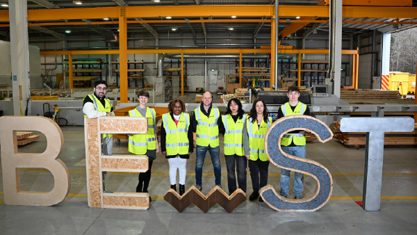 Group of people in high-visibility vests standing in a manufacturing facility behind large letters spelling “BE ST”.