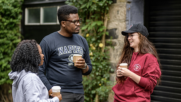 Three students wearing Napier merchandise clothing are chatting whilst holding coffees.