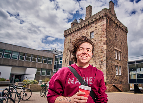 smiling student holding coffee in front of Merchiston's Napier Tower