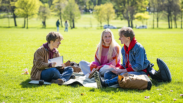 three students with books sit on the grass chatting at The Meadows