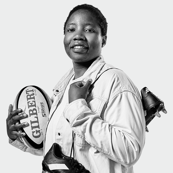 Panashe Muzambe holds her rugby boots and rugby ball, smiling at the camera.